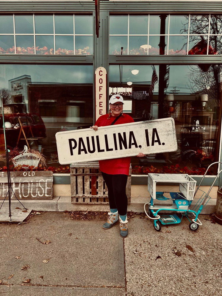 An image of Paulina standing outside with various vintage items on the sidewalk. She's holding a sign which says "Paullina, IA."